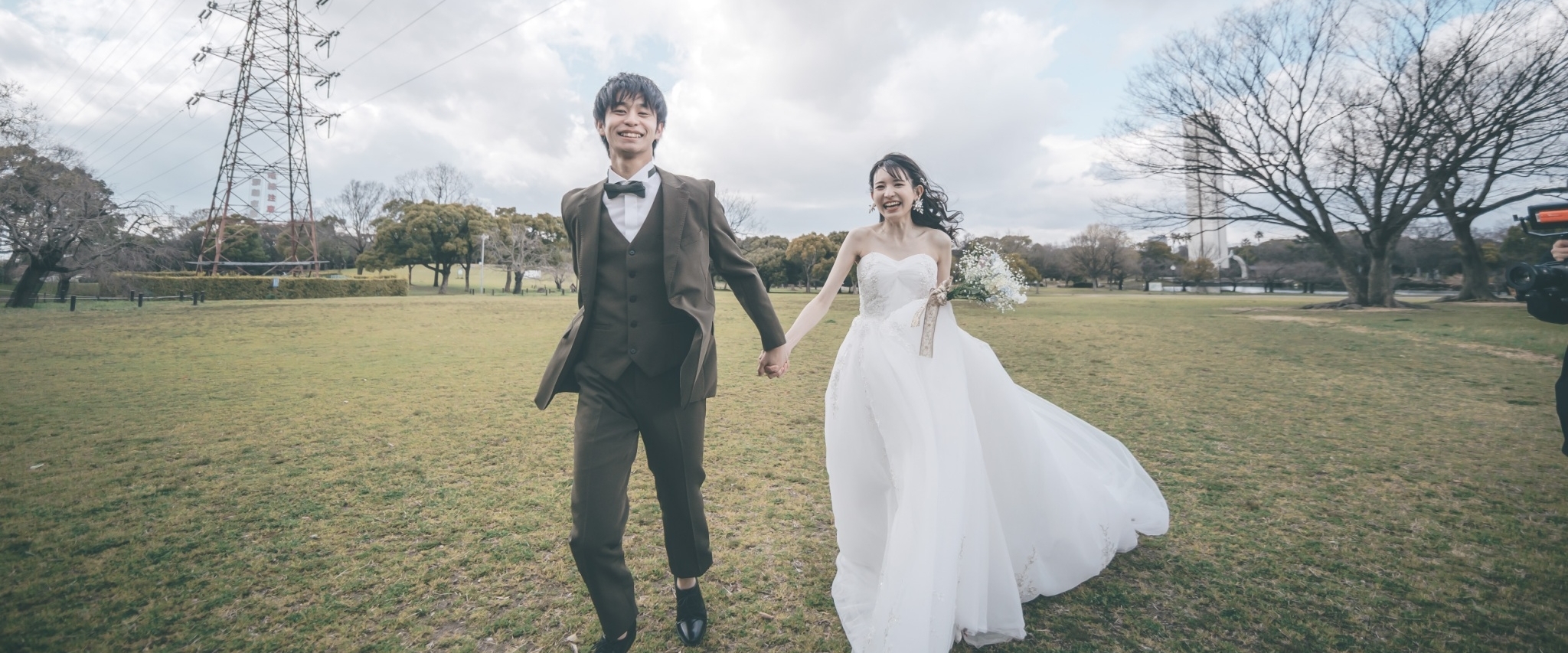 芝生の広場を手をつないで歩く、スリーピースの新郎とウェディングドレスの花嫁。開放感のある洋装ロケーションフォト