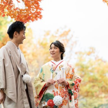 紅葉の下、末広がりの扇子を手に見つめ合う黄色地の花柄着物姿の花嫁と灰茶の着物姿の花婿