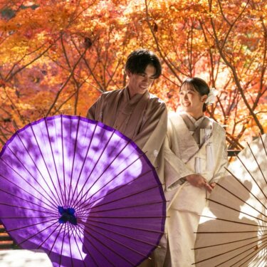燃えるような紅葉を背景に紫と白の和傘を差す、カフェオレ紋付袴の花婿と白無垢姿の花嫁