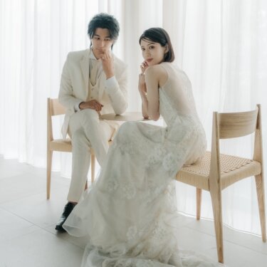 白いカーテンのスタジオで椅子に腰かける白いスーツの新郎とレースドレスの新婦、洋装スタジオフォトの前撮り