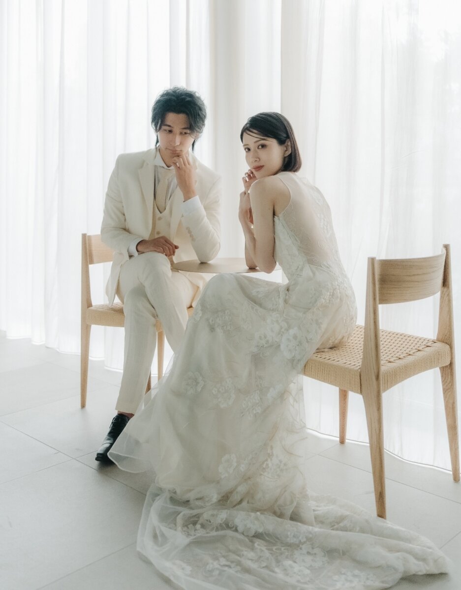 白いカーテンのスタジオで椅子に腰かける白いスーツの新郎とレースドレスの新婦、洋装スタジオフォトの前撮り