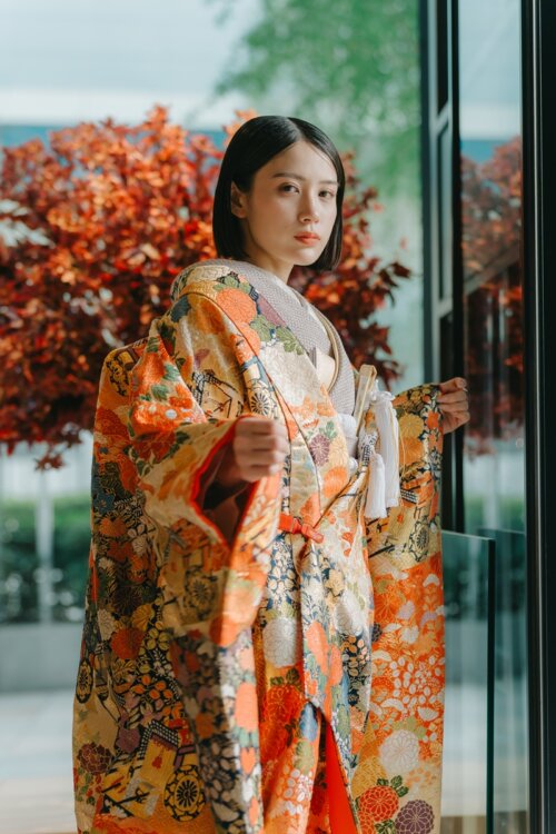 紅葉を背景に佇む、YK16の橙と金地に四季の花々があしらわれた色打掛姿の花嫁