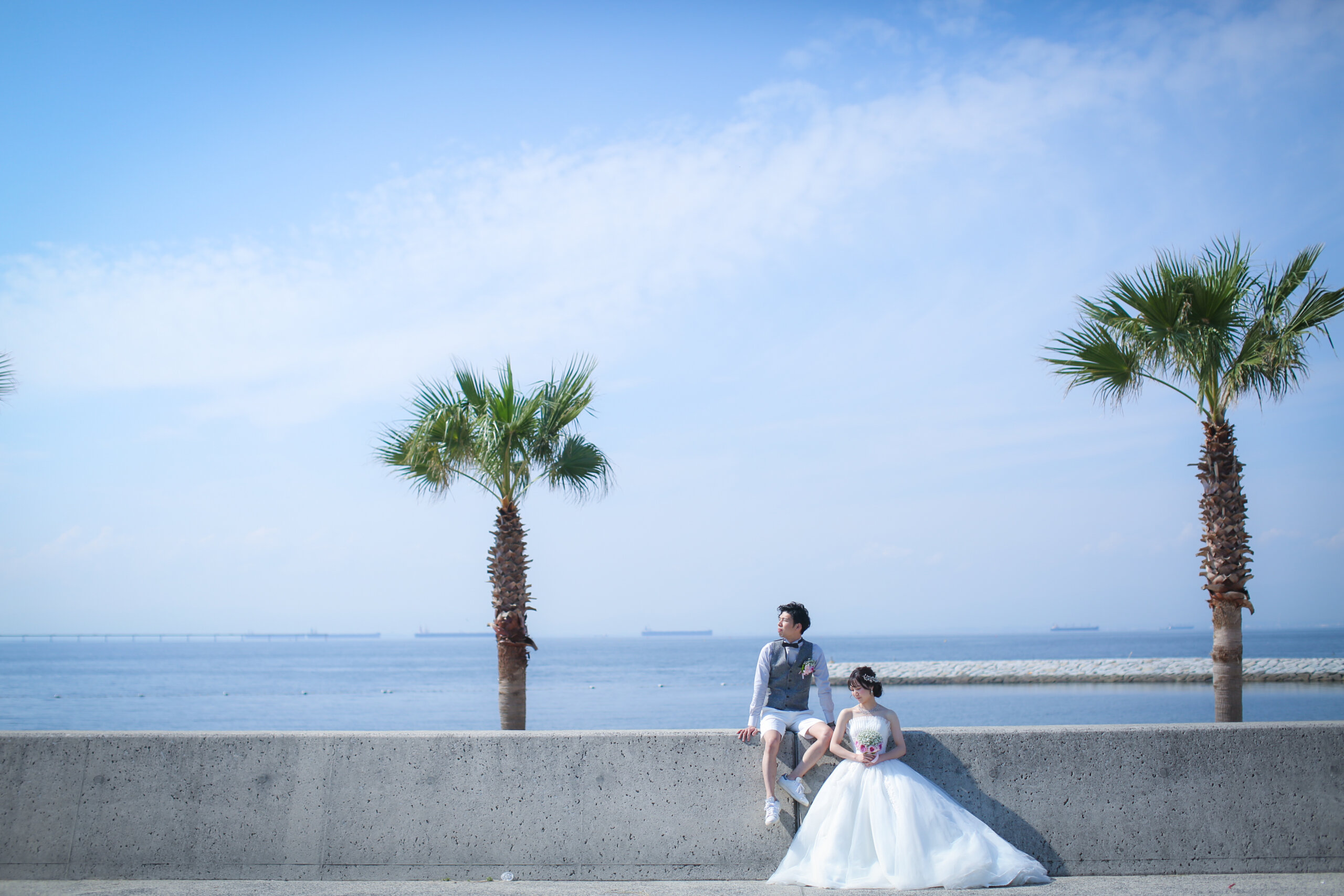 ヤシの木々と海を望む堤防に腰掛ける新郎とブーケを持つウェディングドレスの新婦、りんくうビーチの開放的な眺め