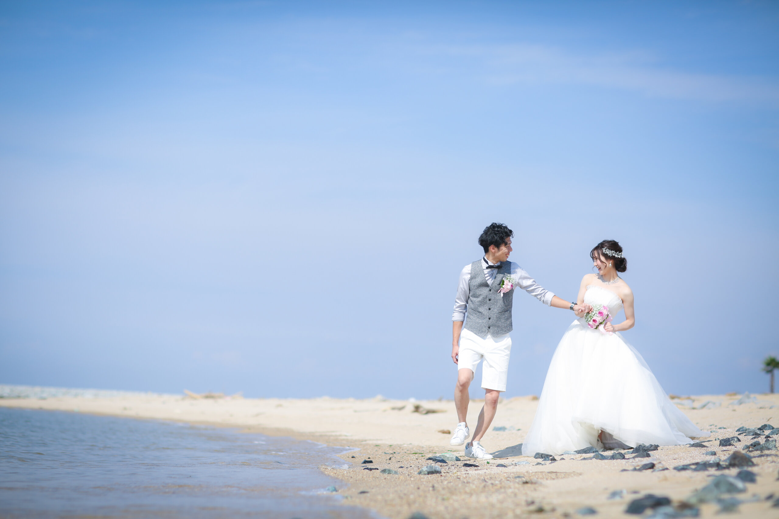 ヤシの木のリゾート感あふれるりんくうビーチを白いショートパンツの新郎とウェディングドレスの新婦が手をつないで歩く