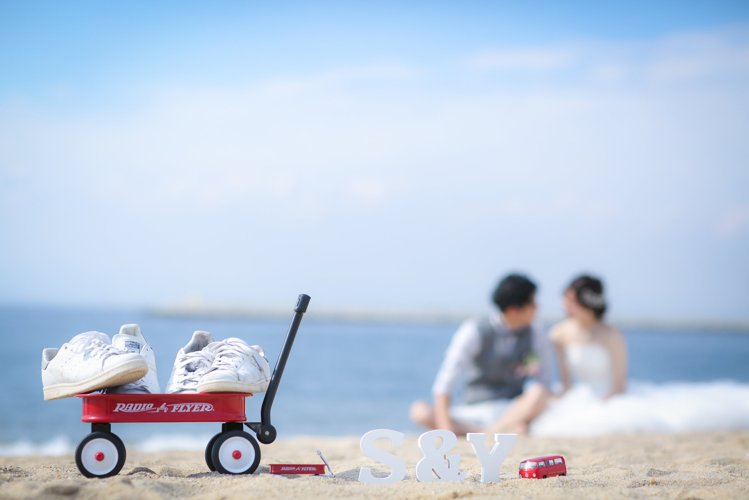 赤いワゴンに乗せた白いスニーカーとイニシャルオブジェ、砂浜でくつろぐ新郎新婦を望むディテールカット