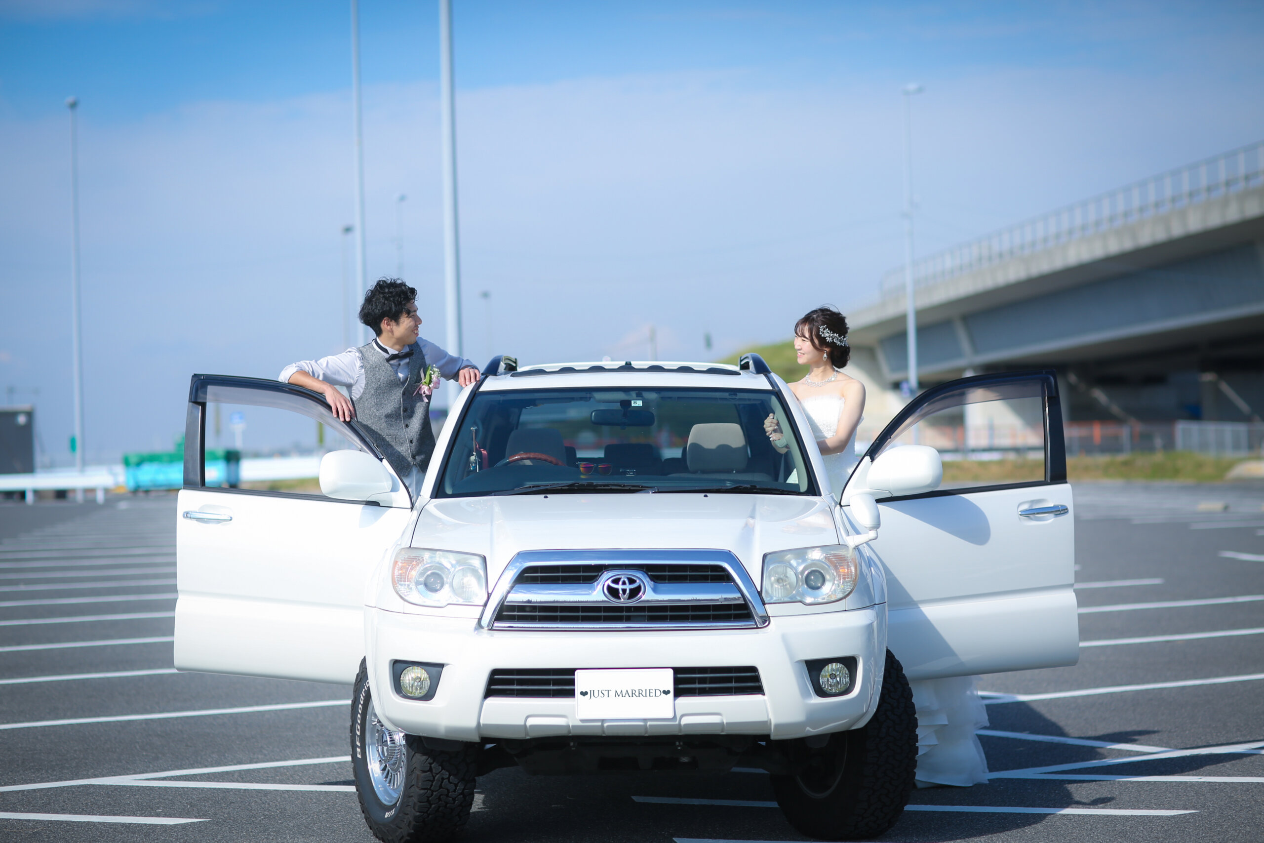「JUST MARRIED」のプレートを付けた白い車のドアを開けて微笑み合う新郎新婦