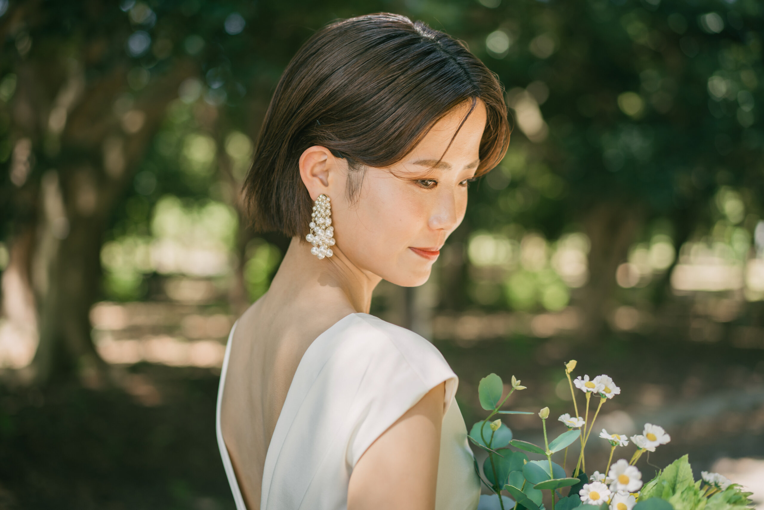木漏れ日の中、白い花のブーケを手に振り返るショートヘアの新婦の横顔