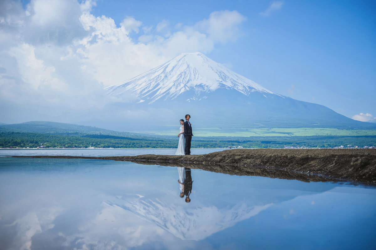 雪化粧の富士山と湖畔に佇むタキシードの新郎とウェディングドレスの新婦、水面に映る富士山の絶景ロケーションフォト