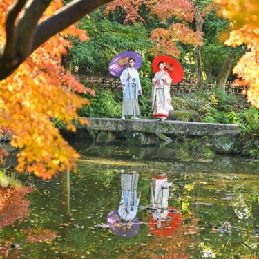 紅葉の木々と池に映る姿を入れて、紫と赤の和傘を差して立つ新郎新婦をとらえた一枚。慶沢園の紅葉で撮る和装前撮りのリフレクション