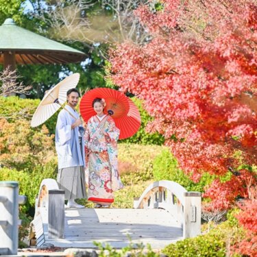 紅葉の庭園の太鼓橋で白い和傘と赤い和傘を差す薄紫の羽織袴の新郎と色打掛の花嫁。秋の日本庭園で撮る和装前撮り