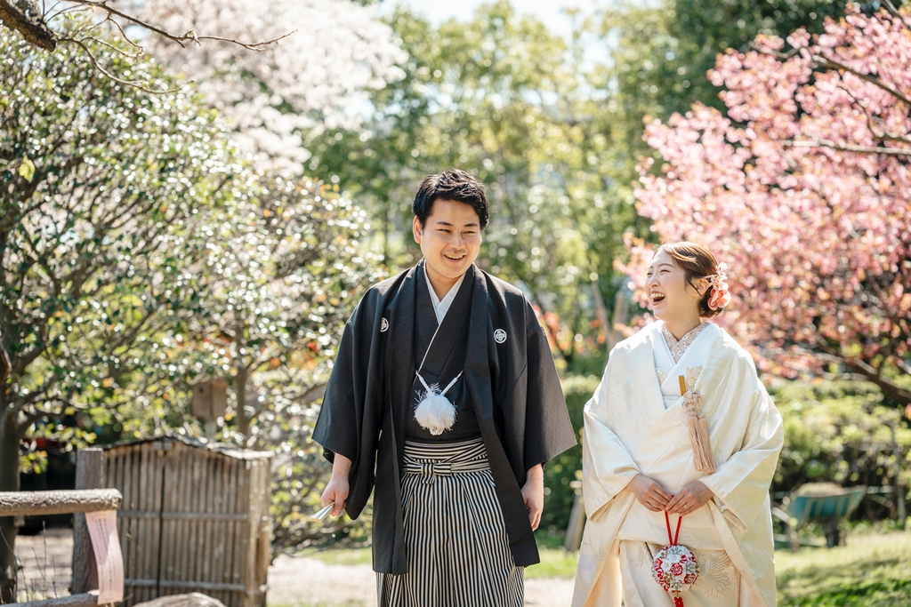 白と桃色の桜に囲まれた小道で笑い合う黒紋付の新郎と白無垢の新婦