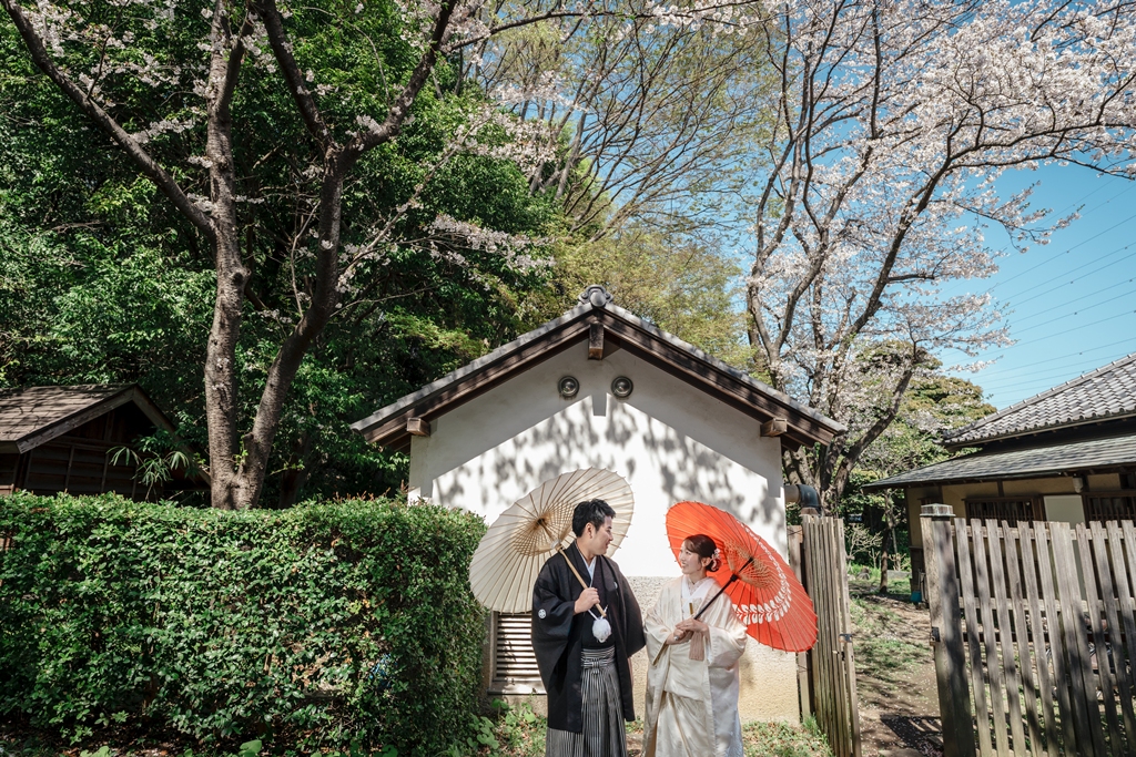 満開の桜を背景に白壁の土蔵の前で白と朱色の和傘を差して見つめ合う黒紋付の新郎と白無垢の新婦