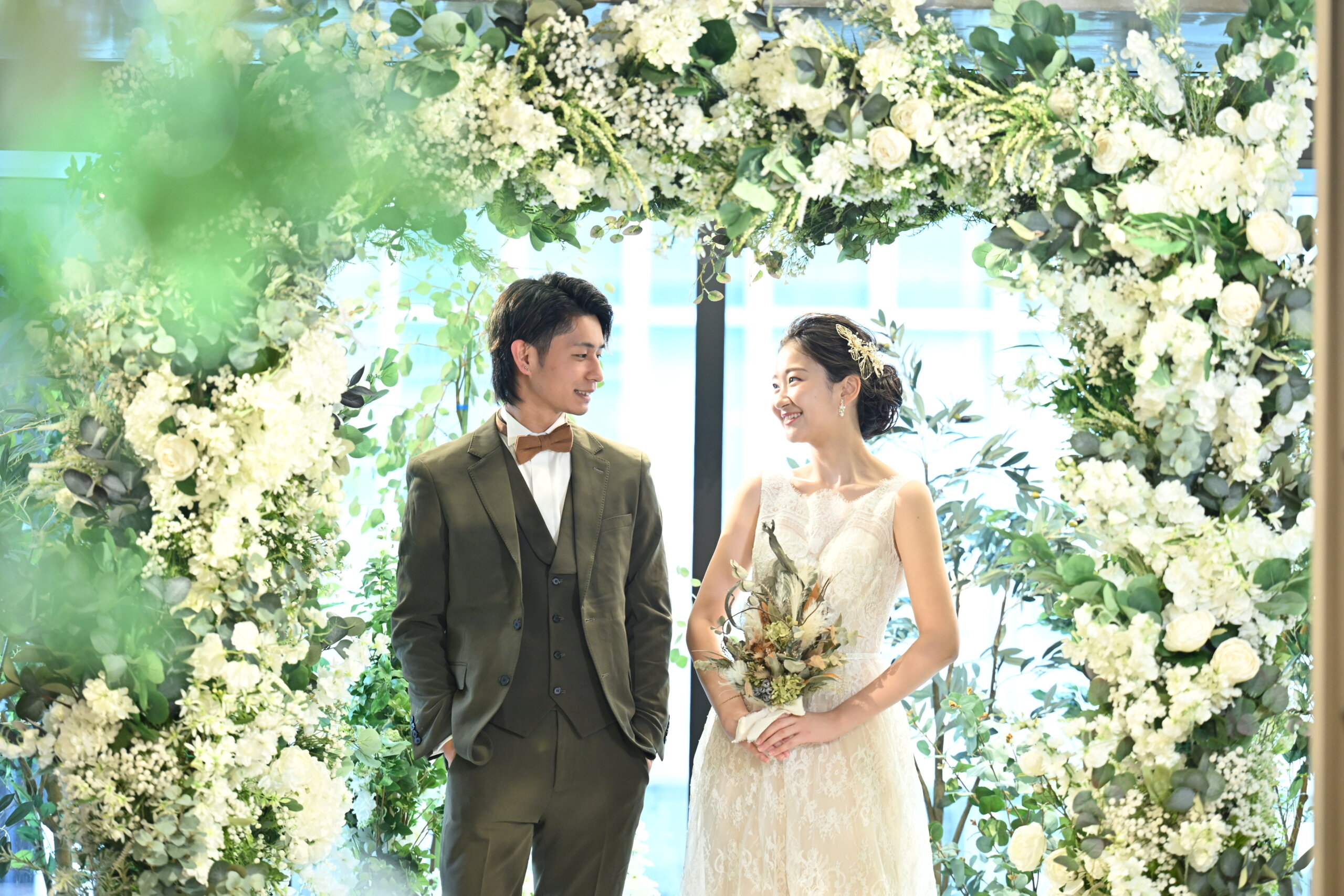 白い花とグリーンのアーチをくぐって見つめ合う、オリーブ色のスリーピース姿の新郎とレースドレス姿の花嫁。心斎橋店ガーデンスタジオでの洋装前撮り