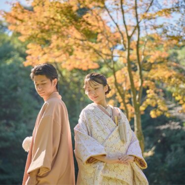 色づいた紅葉を背に背中合わせで微笑む、淡い色打掛の花嫁と紋付の新郎。秋の和装ロケーションフォト