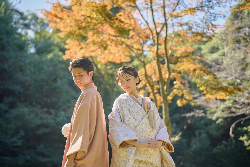 紅葉した木々を背景に背中合わせに立つ薄橙の羽織袴の新郎と花柄の色打掛の新婦