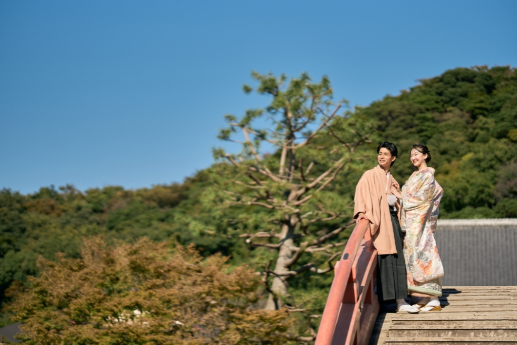 称名寺の反橋の欄干越しに緑豊かな山を望む薄橙の羽織袴の新郎と花柄の着物姿の新婦