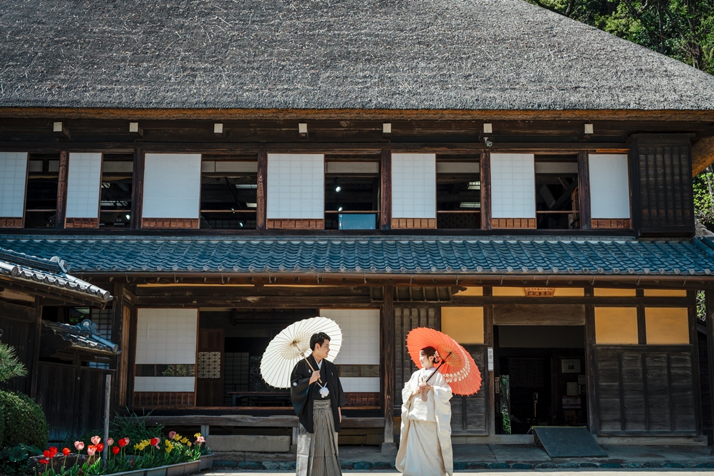 みその公園「横溝屋敷」の茅葺き屋根の古民家を背景に白と朱色の和傘を差して見つめ合う黒紋付の新郎と白無垢の新婦