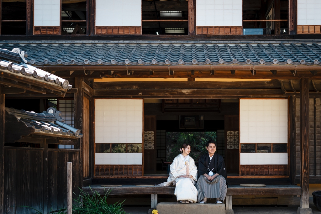横溝屋敷の縁側に並んで腰掛け微笑み合う黒紋付の新郎と白無垢の新婦、重厚な瓦屋根を望む