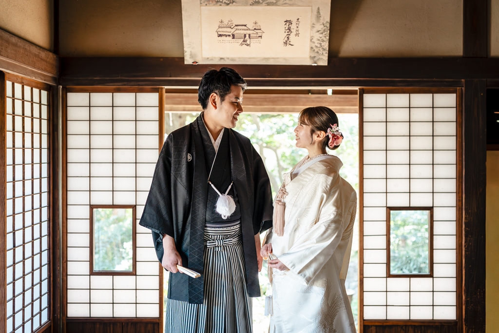 障子の間で見つめ合う黒紋付の新郎と白無垢の新婦、「横溝屋敷」の扁額を掲げた室内