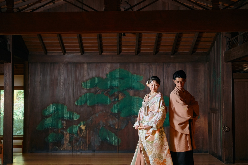 松の絵が描かれた重厚な板戸を背景に佇む花柄の色打掛の新婦と薄橙の羽織袴の新郎、久良岐能舞台の趣ある内観