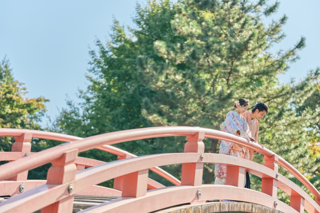 朱塗りの太鼓橋の上で寄り添う花柄の着物姿の新婦と薄橙の羽織袴の新郎