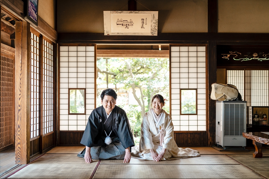 横溝屋敷の座敷で正座して微笑む黒紋付の新郎と白無垢の新婦、庭の緑を望む障子の間