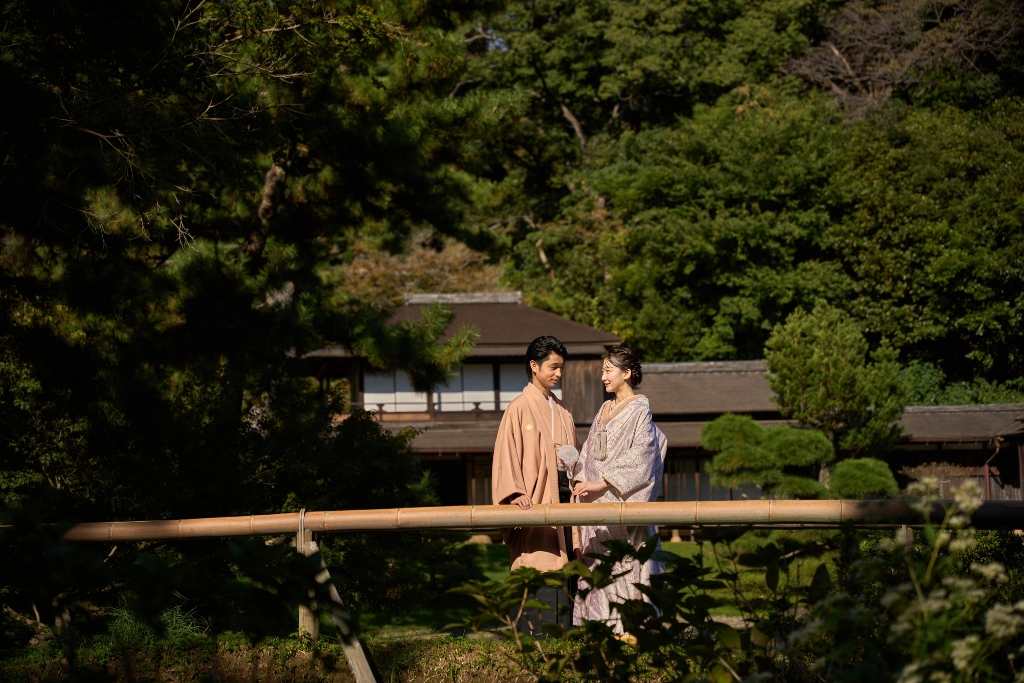 木橋の上で見つめ合う薄橙の羽織袴の新郎と白地の着物姿の新婦、歴史的建造物を望む庭園