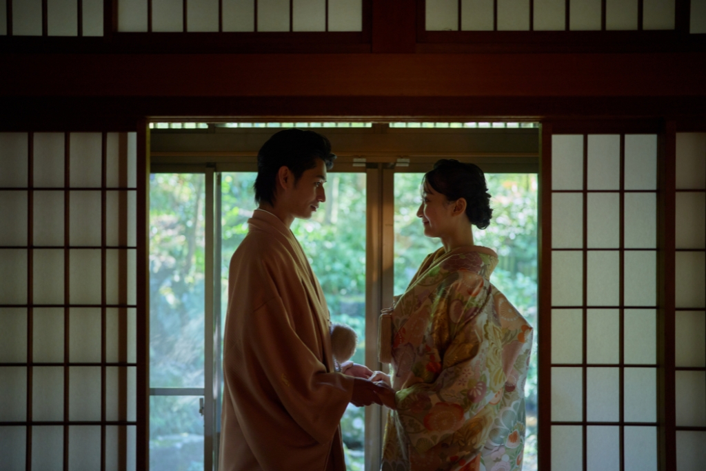 障子の間から緑を望み手を取り合う薄橙の羽織袴の新郎と花柄の色打掛の新婦
