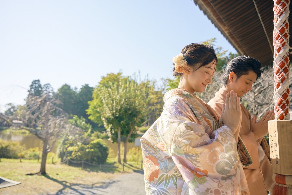 本堂の柱の前で手を合わせる薄橙の羽織袴の新郎と花柄の着物姿の新婦、称名寺の静かな祈りのひととき