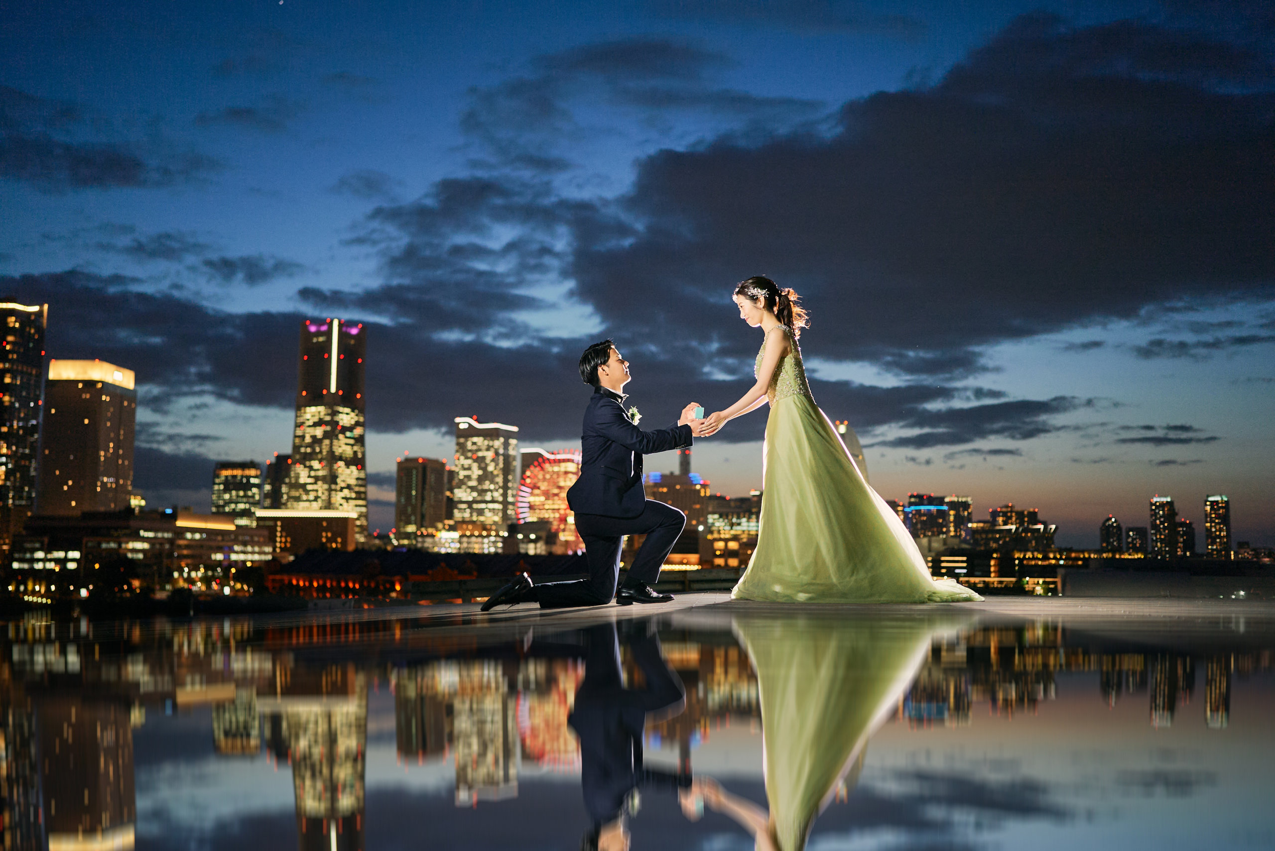 夕暮れのみなとみらいを背景に片膝をついてプロポーズする新郎とグリーンドレスの新婦、特別な瞬間