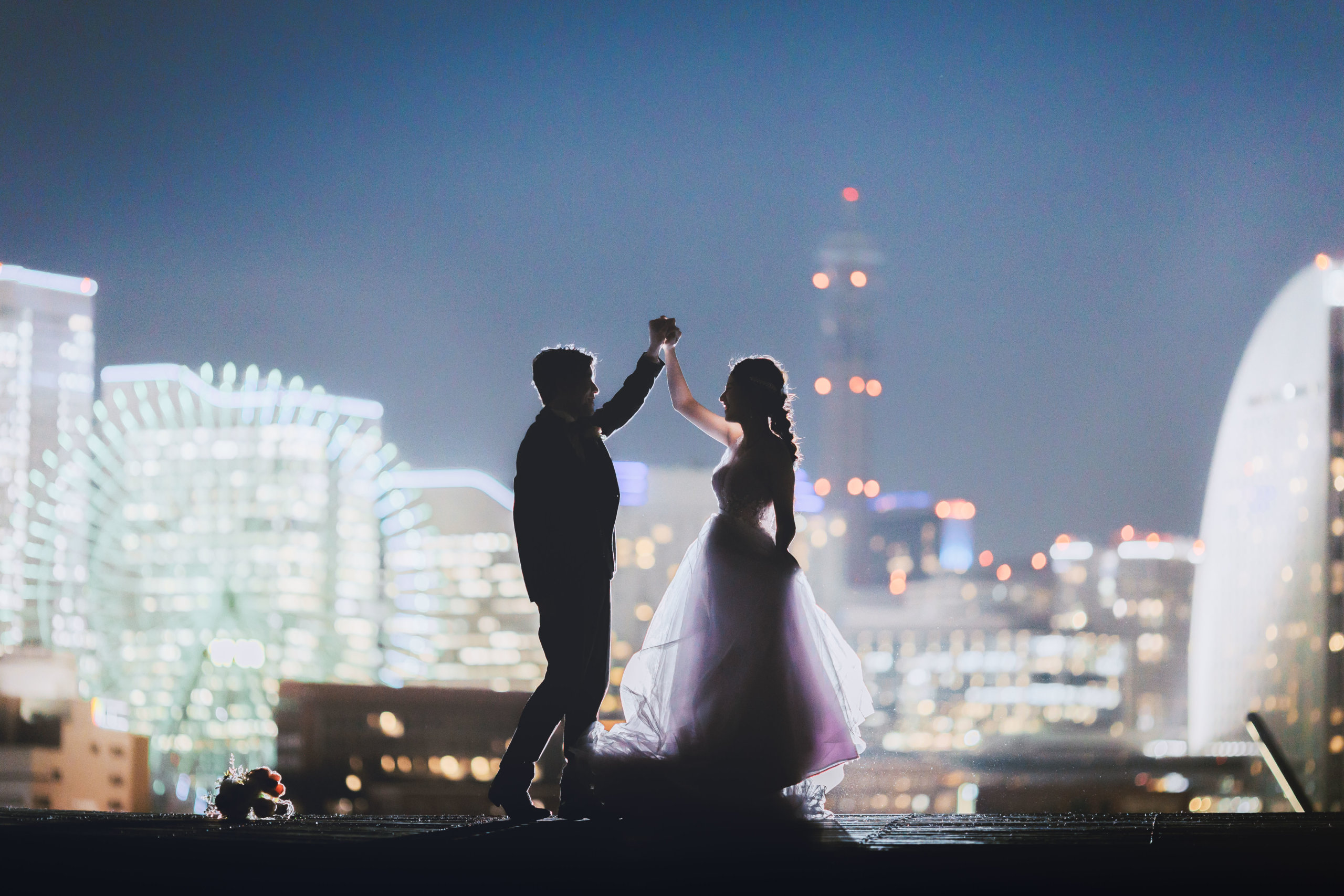 屋上で手を取り合って踊る新郎新婦のシルエット、観覧車とランドマークタワーを望む夜景