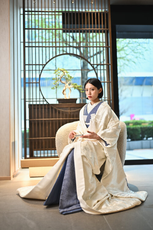 和モダンなラウンジのソファに腰掛け、末広を手にした白無垢姿の花嫁。円窓と盆栽をしつらえた空間での和装前撮り