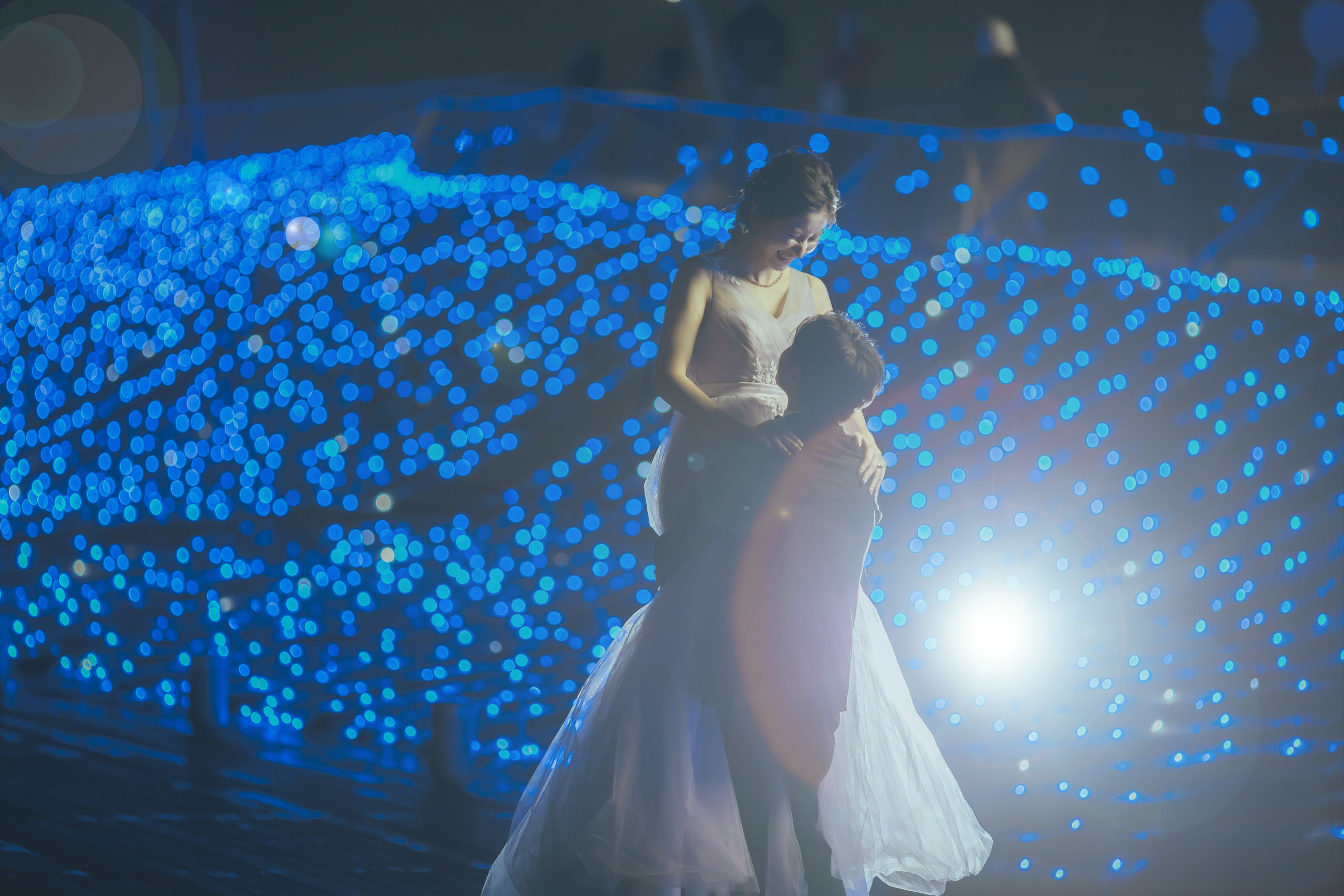 青いイルミネーションの光の中で抱き寄せられる新婦、幻想的な夜のワンショット