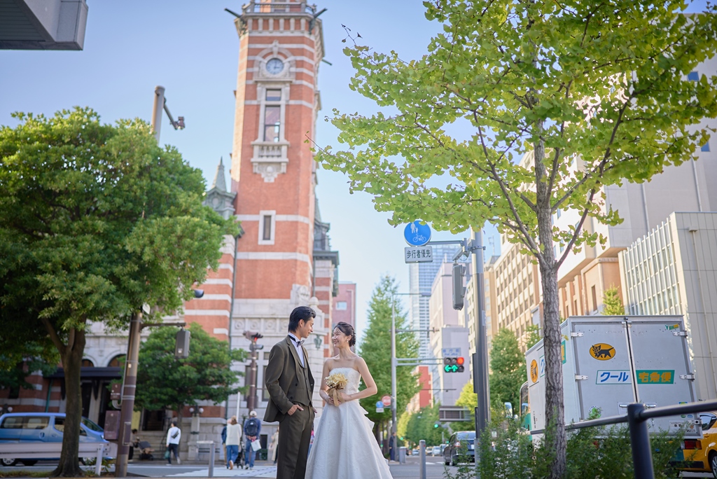 日本大通りの赤レンガ塔を背景にブーケを持つ新婦とブラウンスーツの新郎が見つめ合う、歴史的建造物を望む街並み