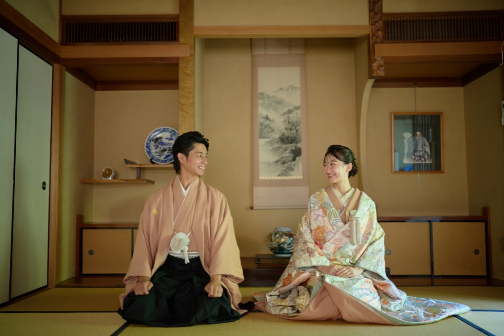 掛け軸を背景に正座して見つめ合う薄橙の羽織袴の新郎と花柄の色打掛の新婦、久良岐能舞台の和室