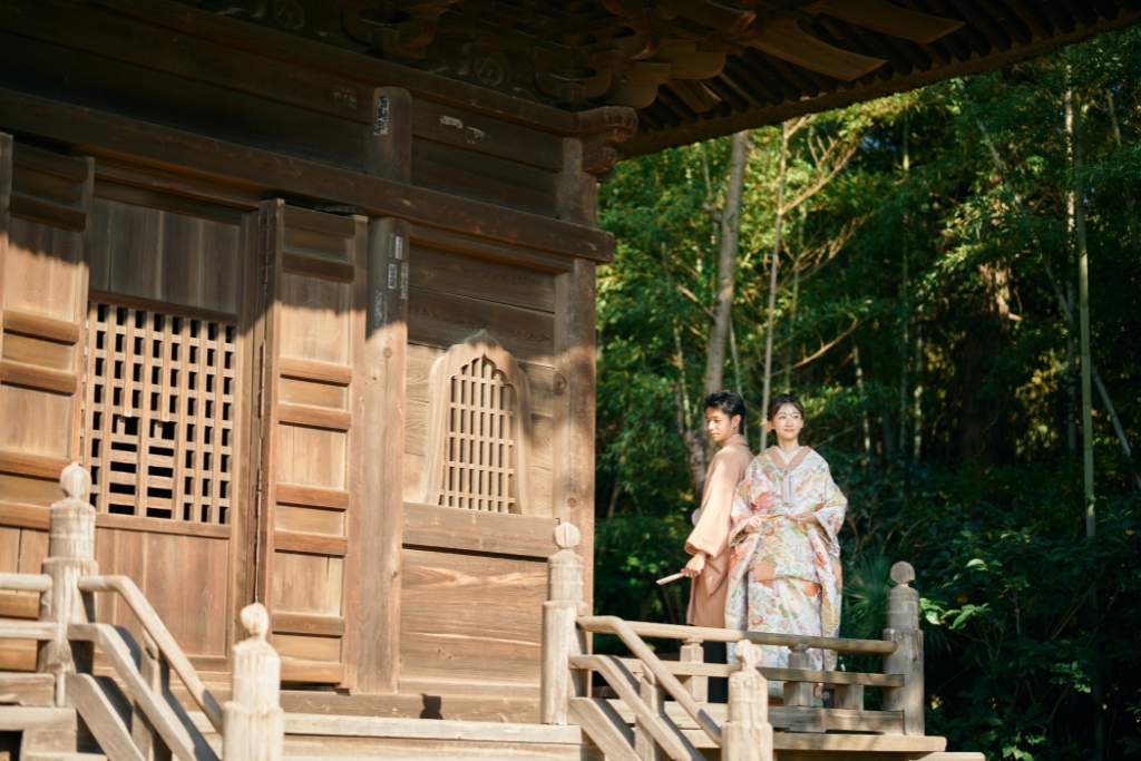 木造の山門を背景に振り返る花柄の着物姿の新婦と薄橙の羽織袴の新郎、竹林を望む静かな境内