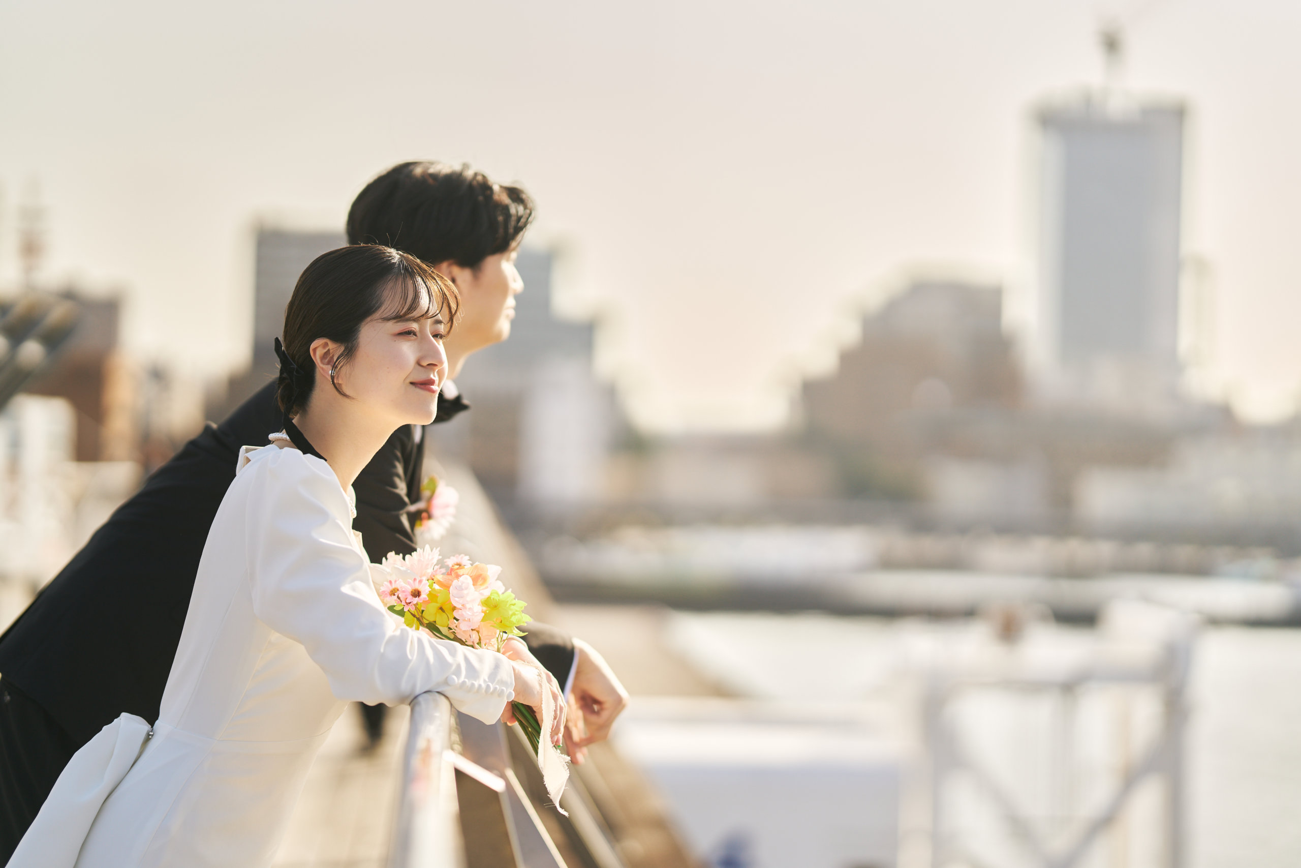 花束を手に寄りかかり横浜の街並みを望む白いドレスの新婦とブラックスーツの新郎
