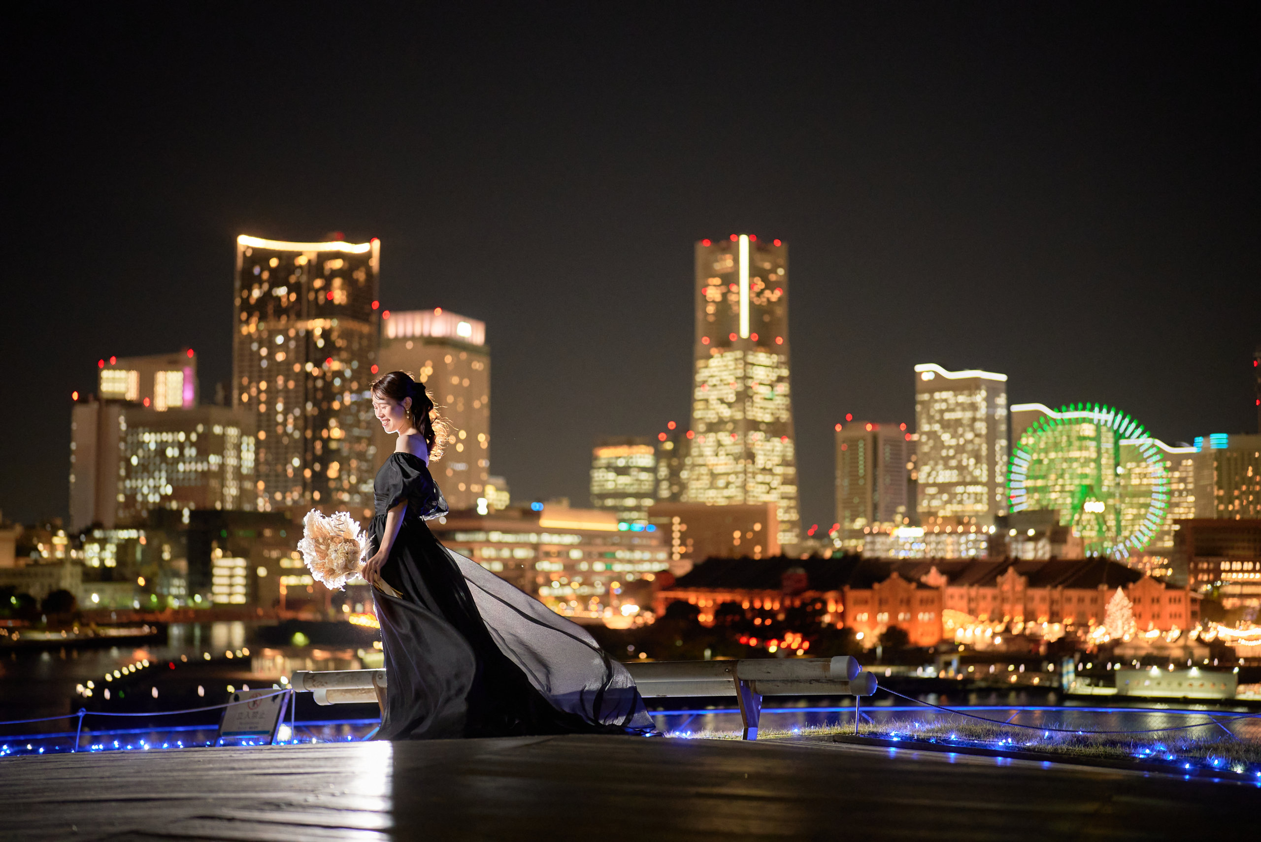 黒いオフショルダードレスの新婦が横浜のみなとみらいの夜景を背景に振り返る、観覧車とランドマークタワーを望む