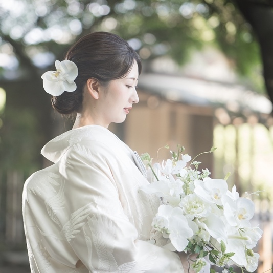白い胡蝶蘭を髪に飾り、ブーケを手に横顔を見せる白無垢姿の花嫁