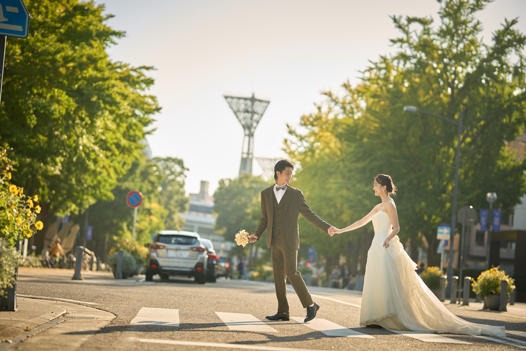 横断歩道を手をつないで渡るブラウンスーツの新郎とウェディングドレスの新婦、街路樹の通りを望む