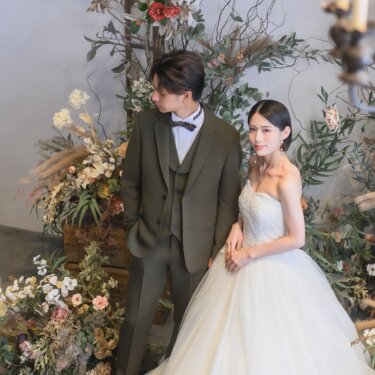 ドライフラワーの装花に囲まれたスタジオで寄り添う新郎新婦。オリーブグリーンのスリーピーススーツとプリンセスラインのウェディングドレスで撮るフォトウェディング