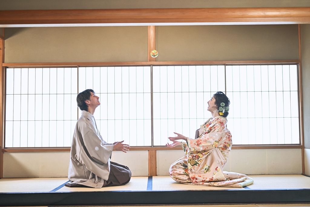 障子の和室で手まりを投げ合い笑い合う、灰茶の羽織と色打掛姿の新郎新婦
