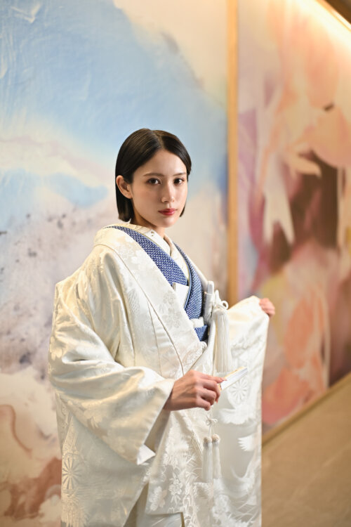 鶴を描いたアートウォールの前で末広を手にした白無垢の花嫁。藍色の掛下を合わせたスタジオ和装フォトウェディング