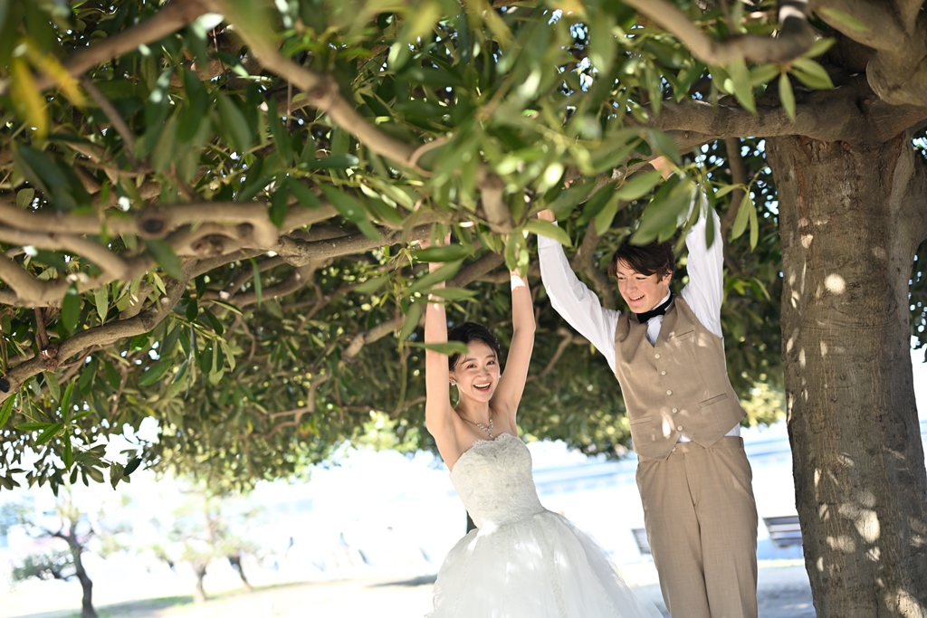 大きな木の枝の下で両手を伸ばしてはしゃぐベストの新郎とウェディングドレスの新婦