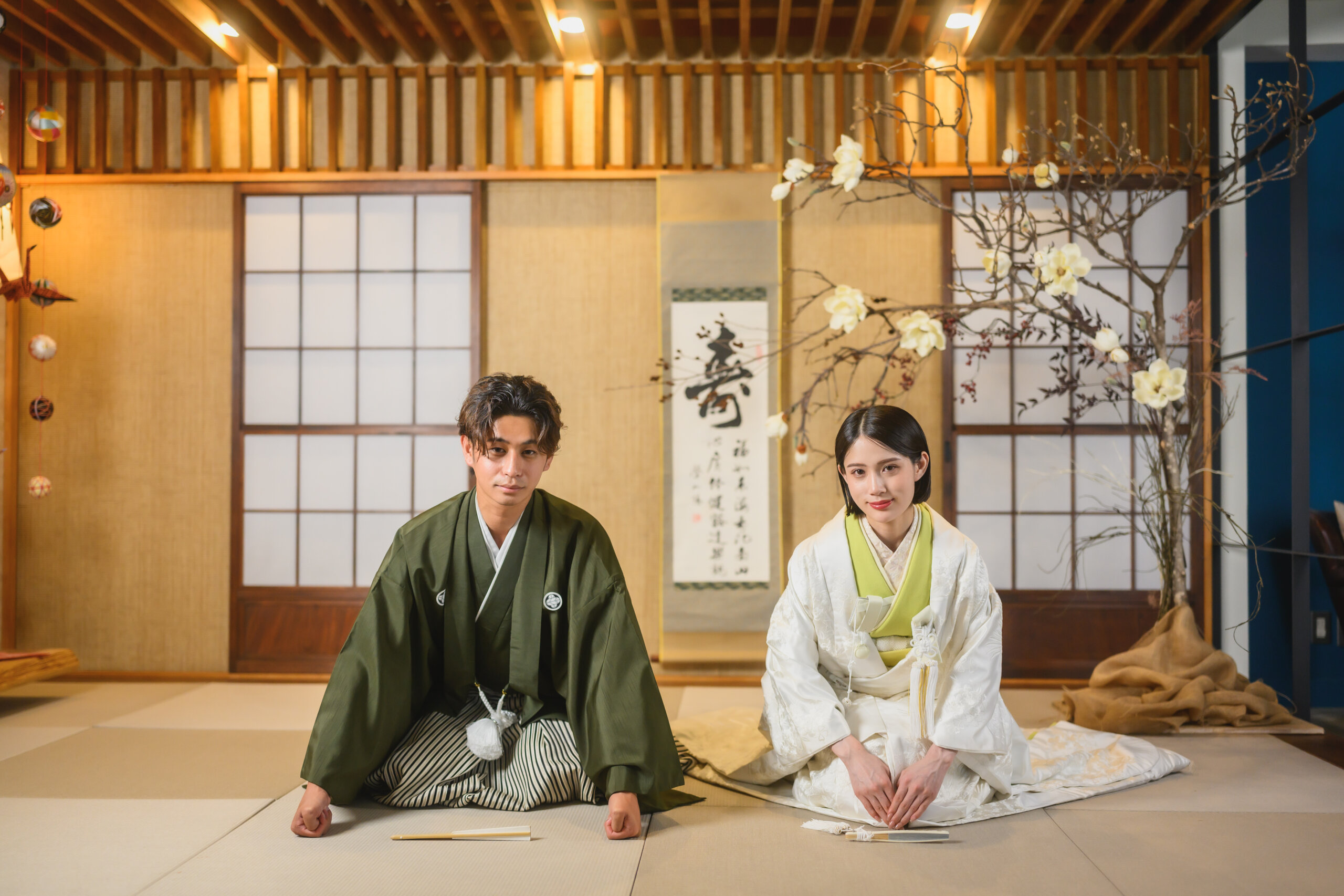 掛け軸と障子のある和室で、畳に正座して並ぶ深緑の羽織に縞袴姿の新郎と白無垢姿の花嫁。THE SKY WEDDING 名古屋のリビングスタジオでの和装前撮り