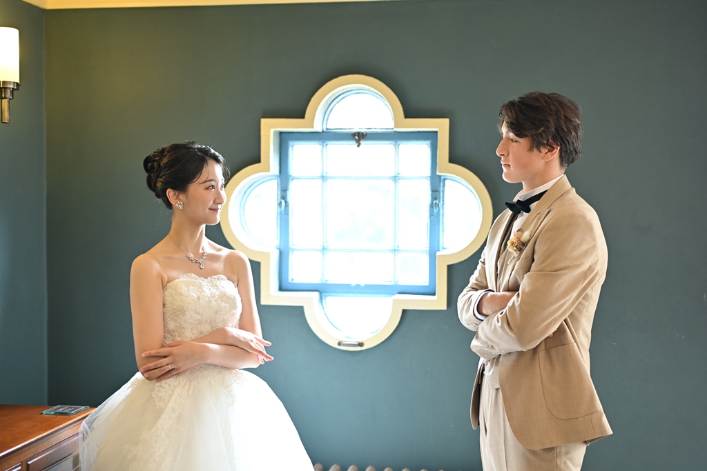 花形のステンドグラス窓を挟んで見つめ合うウェディングドレスの新婦とベージュスーツの新郎