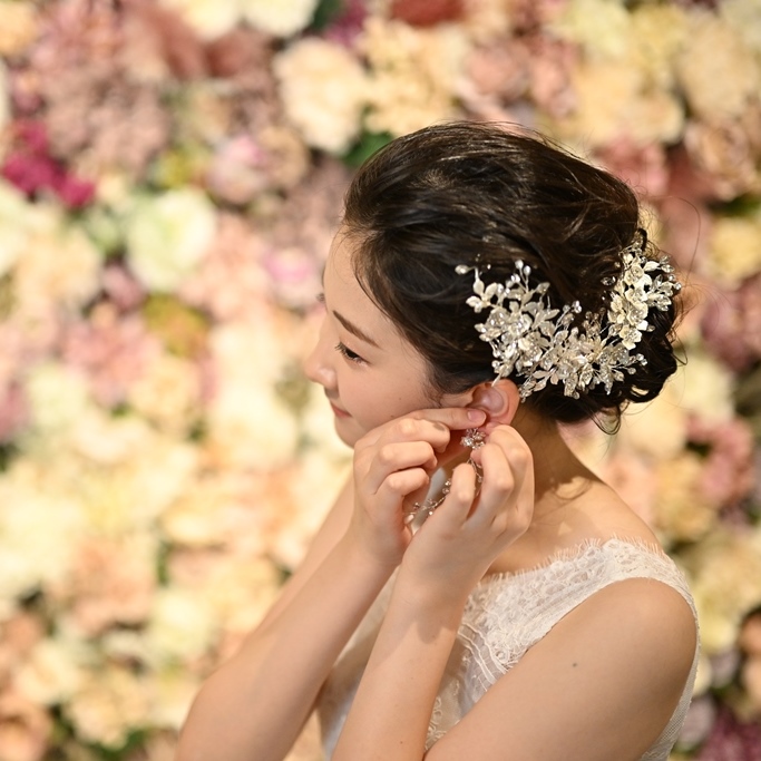 花の壁を背景に、キラキラのビジューヘッドアクセサリーをつけながらピアスを留める花嫁