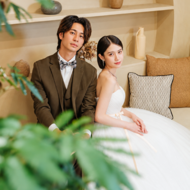 横浜店のスタジオで、ナチュラルな棚とソファの前に並んで座るブラウンのタキシードの新郎とドレスの新婦。手前の緑を生かしたスタジオフォト