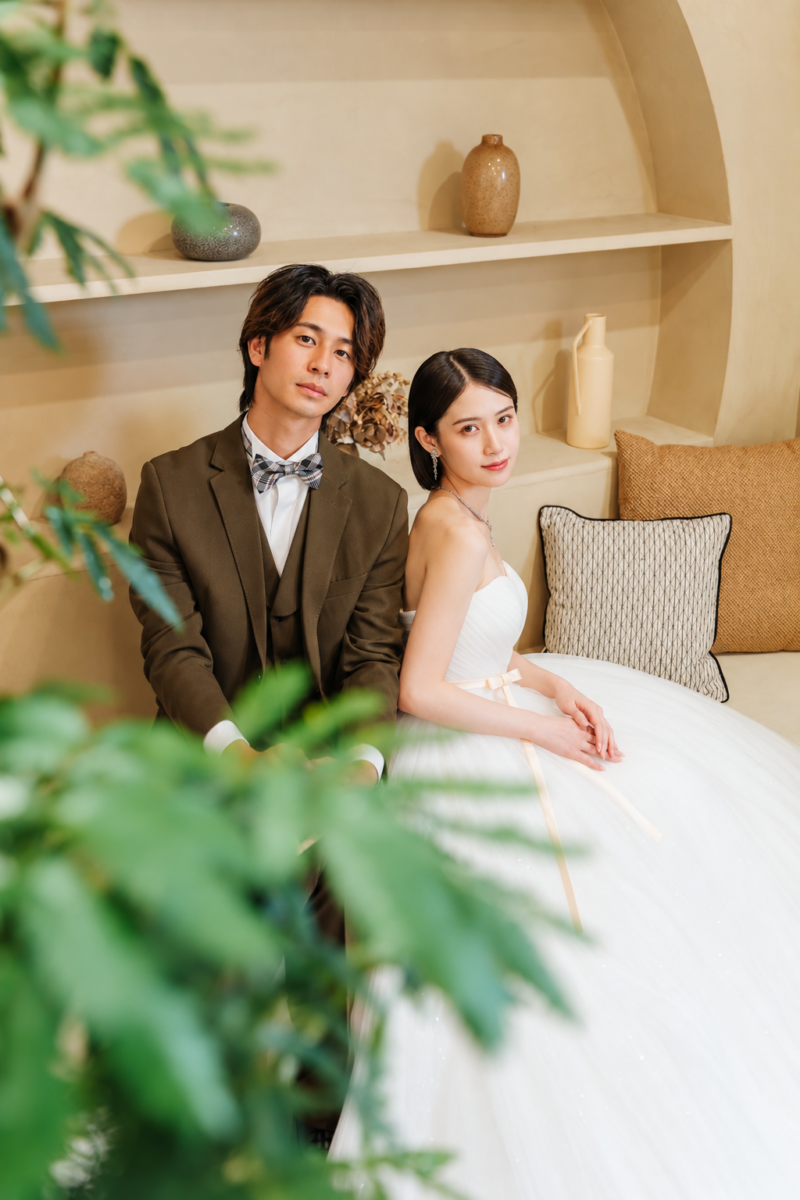 横浜店のスタジオで、ナチュラルな棚とソファの前に並んで座るブラウンのタキシードの新郎とドレスの新婦。手前の緑を生かしたスタジオフォト