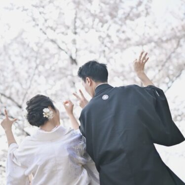 満開の桜を見上げて手でポーズをとる、淡い色の色打掛の花嫁と黒紋付の新郎の後ろ姿。桜で撮る遊び心のある和装前撮り